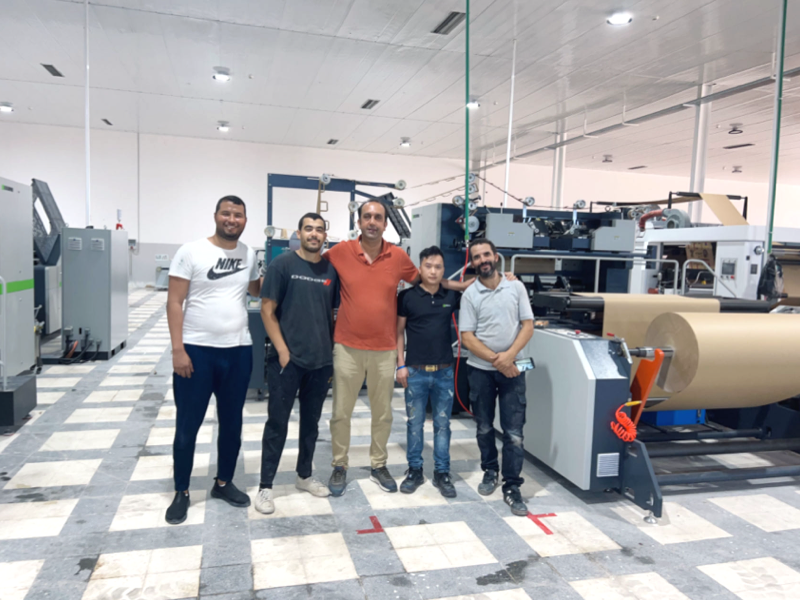 A group of five factory workers standing in a clean industrial facility with machinery
